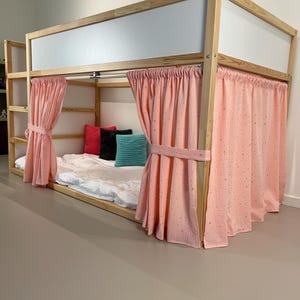May include: A wooden loft bed with a light pink curtain enclosure. The bed frame is natural wood, and the curtains are tied back with matching fabric. The bed has a white mattress and colorful pillows.