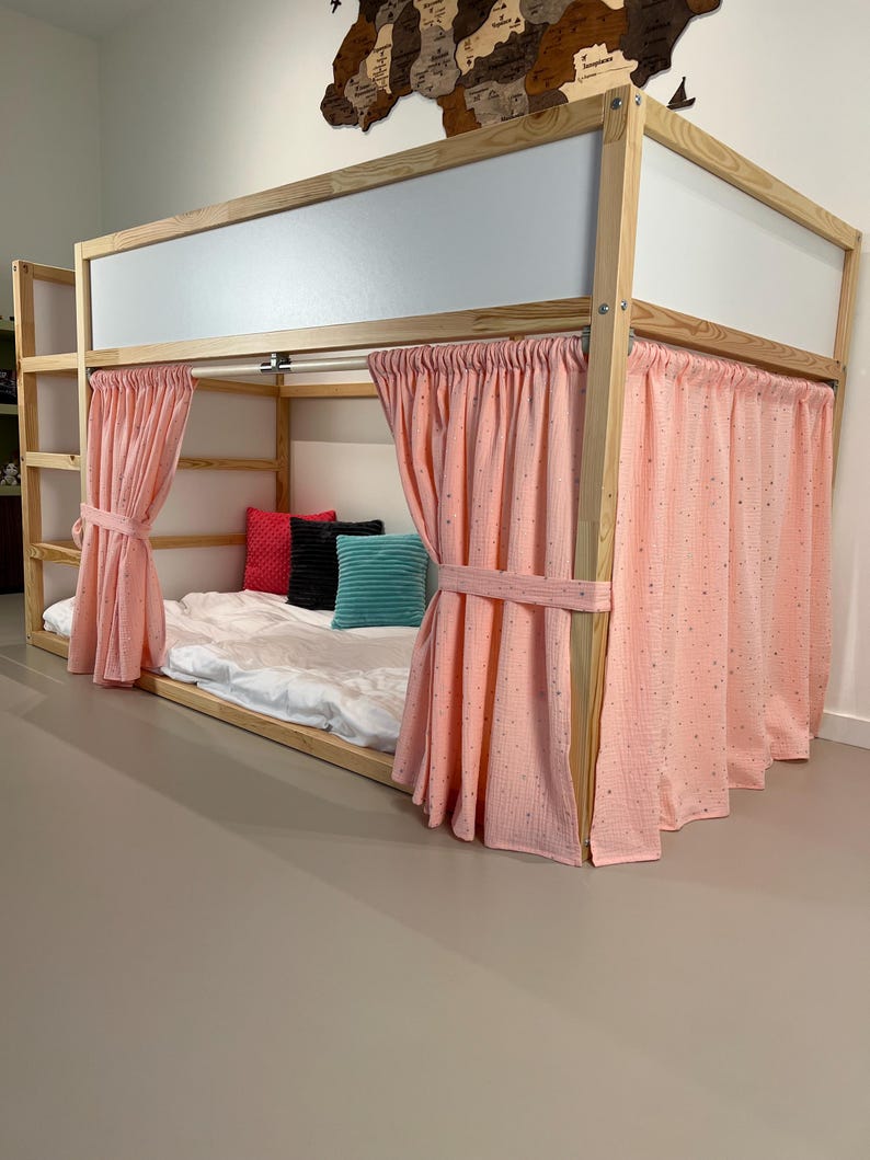 May include: A wooden loft bed with a light pink curtain enclosure. The bed frame is natural wood, and the curtains are tied back with matching fabric. The bed has a white mattress and colorful pillows.