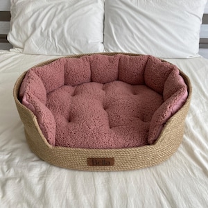 May include: A round pet bed with a woven, natural-colored exterior and a plush, rose-colored interior. The bed has a low profile and a stitched, tufted design. A small, rectangular tag with the name "Bella" is attached to the front.