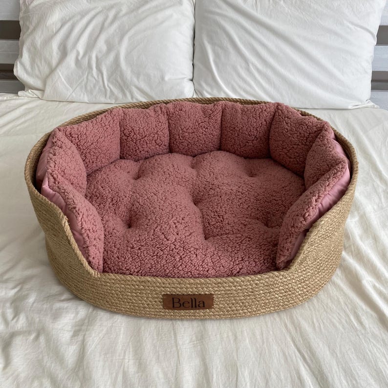 May include: A round pet bed with a woven, natural-colored exterior and a plush, rose-colored interior. The bed has a low profile and a stitched, tufted design. A small, rectangular tag with the name "Bella" is attached to the front.