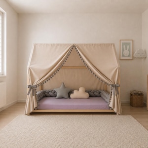 May include: A wooden house-shaped bed frame draped with a beige fabric canopy featuring pom-pom trim. The bed includes a lavender mattress, a gray star-shaped pillow, a cream cloud-shaped pillow, and a gray braided bumper around the perimeter.