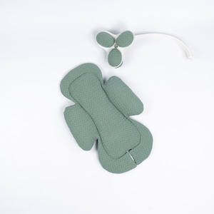 Pode incluir: Um conjunto de artigos para bebé verde sálvia sobre um fundo branco. Os artigos incluem uma almofada de assento acolchoada e um móbile com três elementos em forma de folha. A almofada do assento tem uma superfície texturizada.