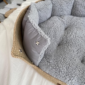 May include: A pet bed with a woven, rope-like border and a soft, gray, plush interior. The bed has a removable, gray, padded cushion with a snap closure. The bed is designed for small pets.