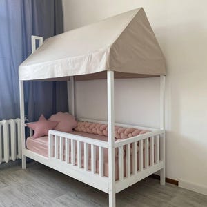 May include: A white wooden house-shaped bed frame with a beige fabric roof canopy. The bed features a slatted side rail, a pink mattress, a pink braided bumper, and two pink decorative pillows in the shape of a star and a cloud.