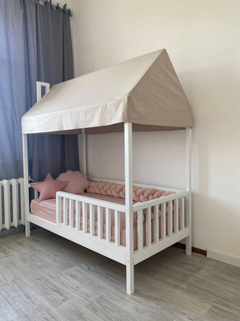 May include: A white wooden house-shaped bed frame with a beige fabric roof canopy. The bed features a slatted side rail, a pink mattress, a pink braided bumper, and two pink decorative pillows in the shape of a star and a cloud.