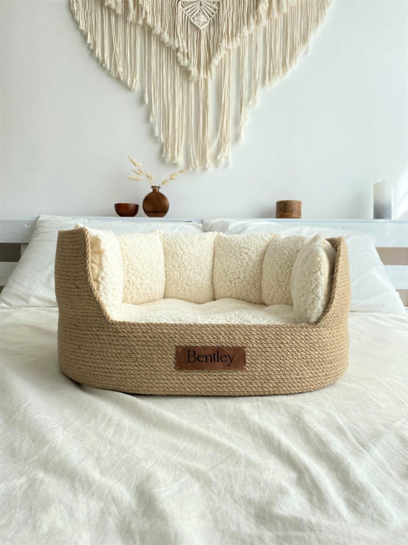 May include: A beige woven dog bed with a soft, cream-colored interior and bolster pillows. The bed has a rectangular shape with rounded edges and a brown leather tag that reads "Bentley".