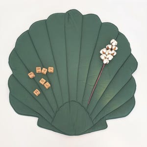 Könnte beinhalten: Eine dunkelgrüne, muschelförmige Spielmatte mit gesteppten Details. Die Matte hat einen gewellten Rand und ist mit Holzbuchstabenblöcken und einem Baumwollzweig verziert. Die Matte befindet sich auf einem weißen Hintergrund.