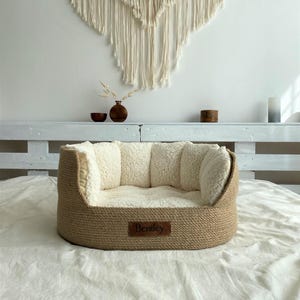 May include: A beige pet bed with a woven exterior and a soft, cream-colored interior. The bed has a rectangular shape with raised sides and a small, rectangular tag that reads "Bentley". The bed is placed on a white sheet.