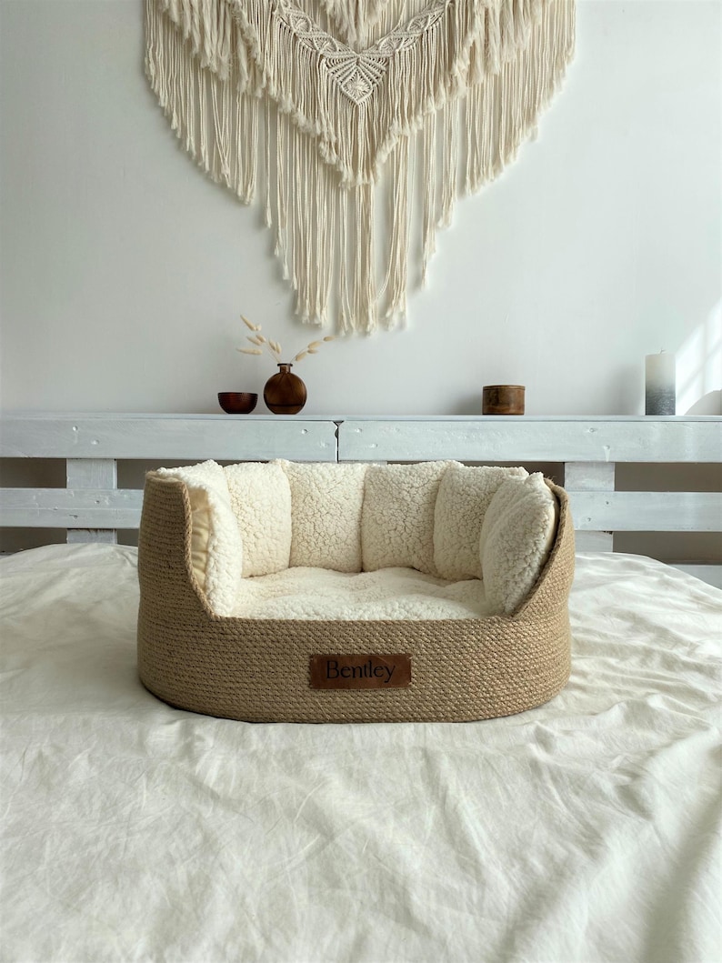 May include: A beige pet bed with a woven exterior and a soft, cream-colored interior. The bed has a rectangular shape with raised sides and a small, rectangular tag that reads "Bentley". The bed is placed on a white sheet.