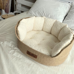 May include: Oval pet bed with a woven, tan-colored exterior and a plush, cream-colored interior. The bed has a small, brown leather tag that reads "Bentley". The bed is placed on a white bedspread.