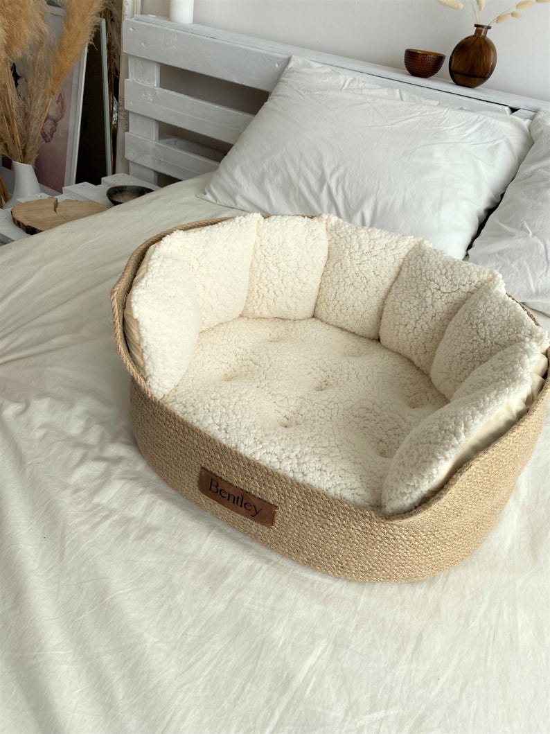 May include: Oval pet bed with a woven, tan-colored exterior and a plush, cream-colored interior. The bed has a small, brown leather tag that reads "Bentley". The bed is placed on a white bedspread.