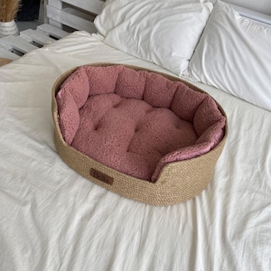 May include: An oval pet bed with a woven jute exterior and a plush, pink interior. The bed features a soft, cushioned center and a raised edge for comfort. A small, brown label with the name "Bella" is attached to the front.