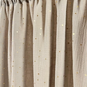 Pode incluir: Cortinas bege adornadas com estrelas e pontos dourados. O tecido &eacute; franzido na parte superior, criando dobras verticais. As estrelas douradas s&atilde;o distribu&iacute;das uniformemente pelos pain&eacute;is, adicionando um toque de fantasia ao fundo neutro.