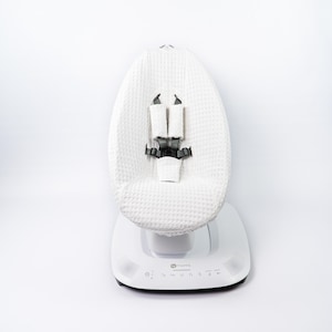 Op de afbeelding: Een witte babyschommel met een gestructureerde, wafelgeweven zitting en een witte basis. De zitting heeft grijze veiligheidsriempjes. De basis heeft bedieningsknoppen en luidsprekers.