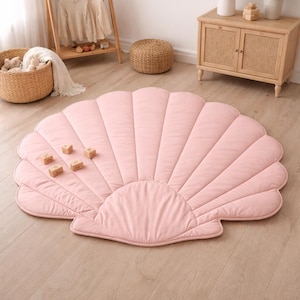 May include: A large, pink, seashell-shaped play mat with a quilted design. The mat is on a light wood floor. Several wooden blocks are on the mat. The mat is in a room with a wooden cabinet, a small woven stool, and a clothes rack.