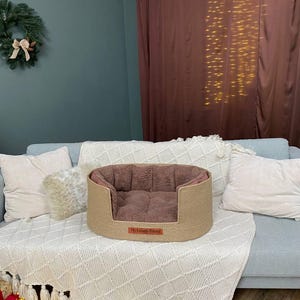 May include: A beige dog bed with a brown interior sits on a light blue couch. The bed has a woven exterior and a soft, plush interior. A small rectangular tag on the front of the bed reads "My Lovely Friend".