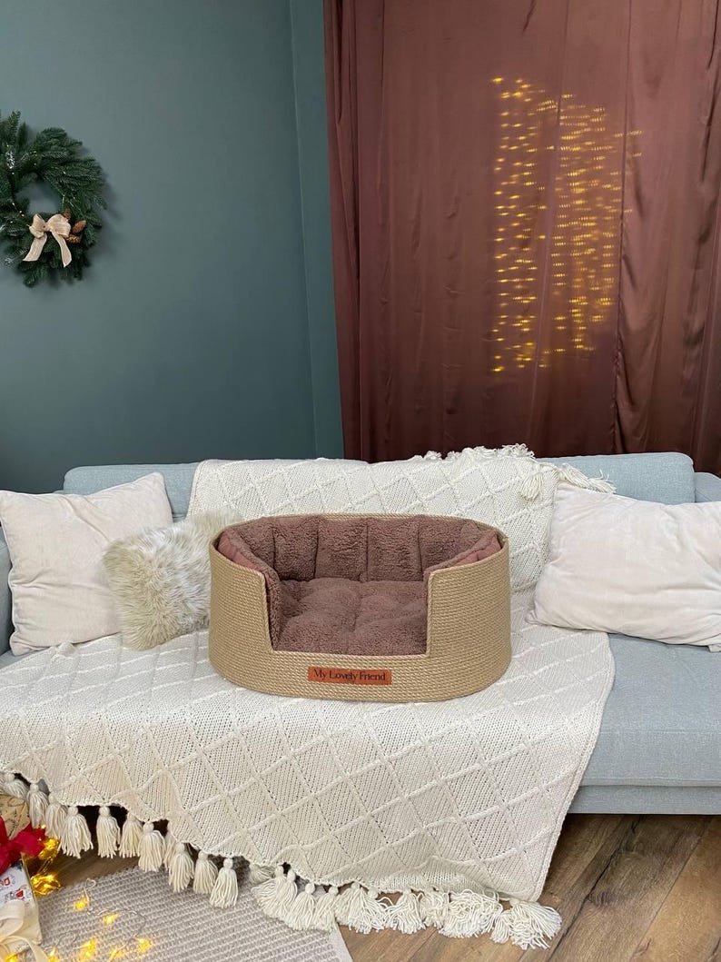 May include: A beige dog bed with a brown interior sits on a light blue couch. The bed has a woven exterior and a soft, plush interior. A small rectangular tag on the front of the bed reads "My Lovely Friend".