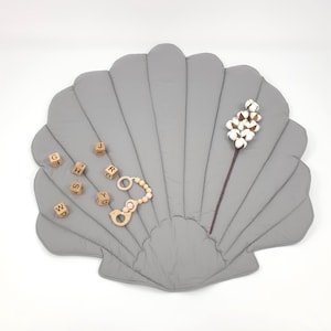 Könnte beinhalten: Eine graue, muschelförmige Spielmatte mit Steppdesign. Buchstabenblöcke aus Holz und ein Beißring aus Holz mit rosa Perlen sind auf der Matte platziert. Ein Zweig Baumwolle sorgt für eine natürliche Note.
