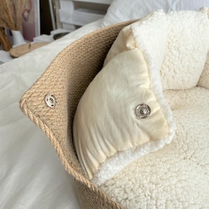 May include: A pet bed with a woven jute exterior and a soft, cream-colored interior. The bed features a removable, plush cushion and a snap closure. The overall design is neutral and cozy, suitable for small pets.