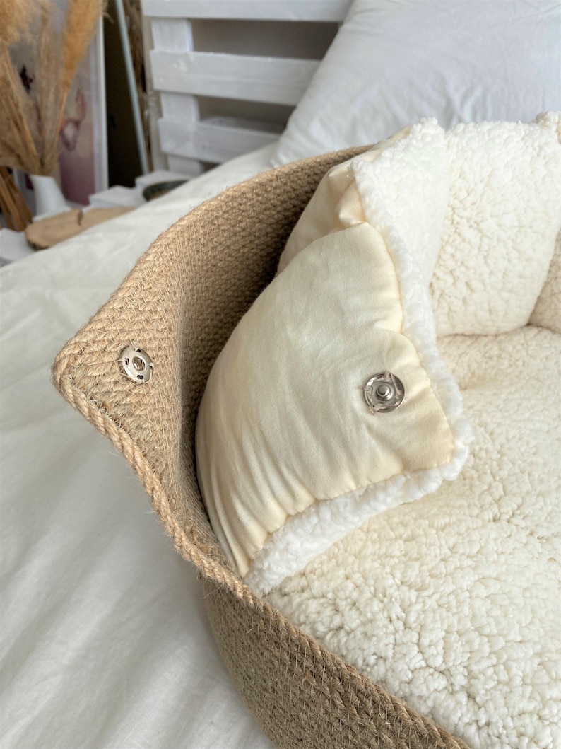 May include: A pet bed with a woven jute exterior and a soft, cream-colored interior. The bed features a removable, plush cushion and a snap closure. The overall design is neutral and cozy, suitable for small pets.