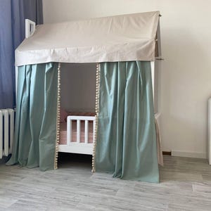 May include: A white house-shaped bed frame featuring a beige fabric roof canopy and sage green curtains with cream-colored pom-pom trim along the inner edges. The bed includes a white wooden safety rail and a light pink mattress cover inside.