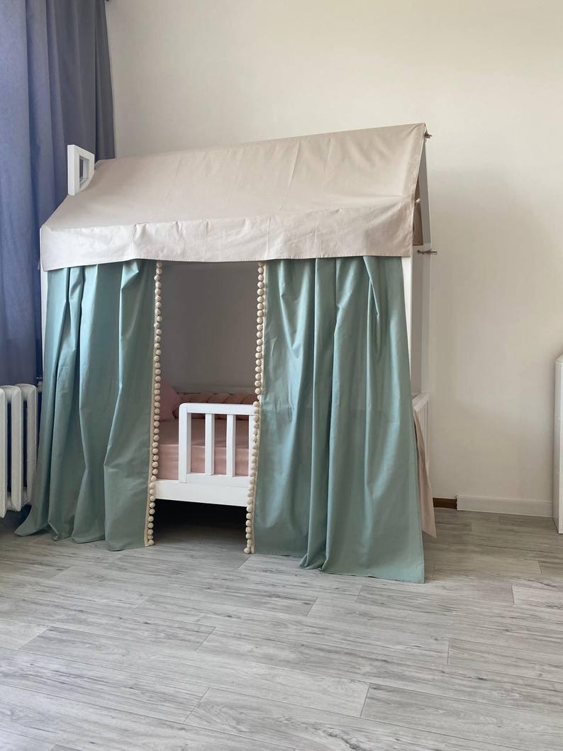 May include: A white house-shaped bed frame featuring a beige fabric roof canopy and sage green curtains with cream-colored pom-pom trim along the inner edges. The bed includes a white wooden safety rail and a light pink mattress cover inside.
