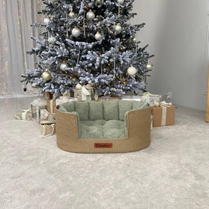 May include: A woven dog bed with a light green cushion and a brown leather tag that reads "Charlee." The bed is oval-shaped and sits on a light gray carpet. A decorated Christmas tree is in the background.
