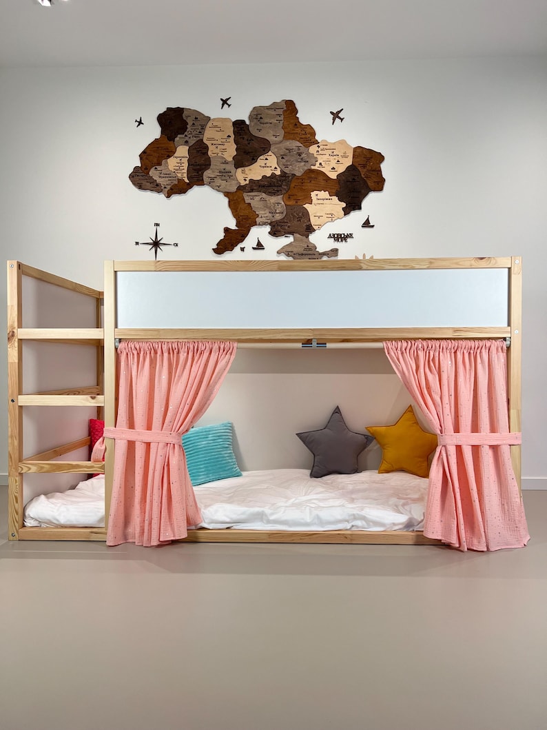 Pode incluir: Uma cama de beliche de madeira com estrutura de madeira clara, cortinas rosa e almofadas em forma de estrela. Um mapa de madeira da Ucr&acirc;nia est&aacute; pendurado na parede acima da cama. A cama tem uma escada para acesso &agrave; cama superior.