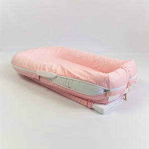 Puede incluir: Un cojín para bebé rosa con cremallera y ribete blancos. El cojín es rectangular con bordes redondeados y un interior acolchado elevado. Diseñado para la comodidad y seguridad de los bebés.