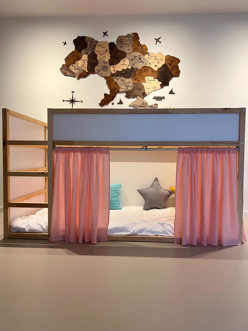 May include: A wooden bunk bed with a light-colored frame and pink curtains. The bed has a built-in ladder and shelves. A wooden map of Ukraine hangs on the wall above the bed, with a compass and airplane cutouts.