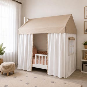May include: A white house-shaped bed frame featuring a beige fabric roof, white curtains with pom-pom trim, and a side window with a roll-up shade. The bed includes a white safety rail and is dressed with a solid peach-colored mattress and pillow.