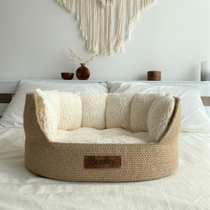May include: A round dog bed with a woven, light brown exterior and a soft, cream-colored interior. The bed has a rectangular leather tag that reads "Bentley". The bed is designed for comfort and support.