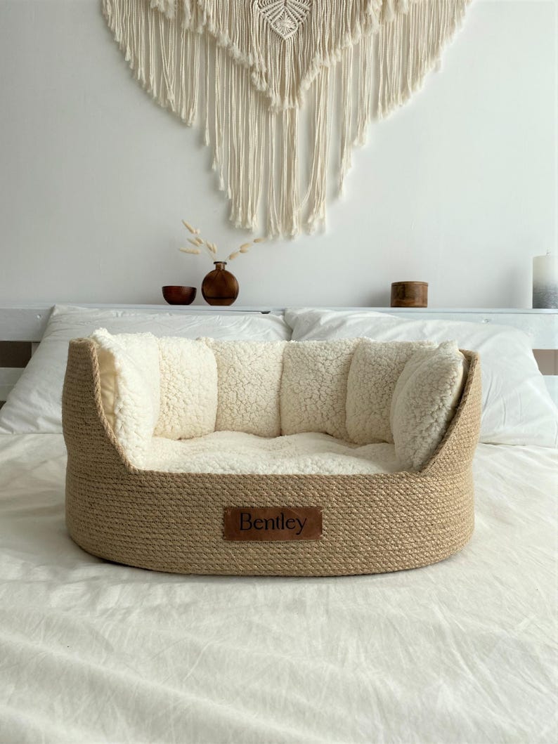 May include: A round dog bed with a woven, light brown exterior and a soft, cream-colored interior. The bed has a rectangular leather tag that reads "Bentley". The bed is designed for comfort and support.