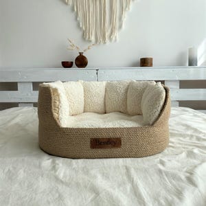 May include: A dog bed with a woven, light brown exterior and a soft, cream-colored interior. The bed has a rectangular shape with raised sides and a small, brown leather tag that reads "Bentley".