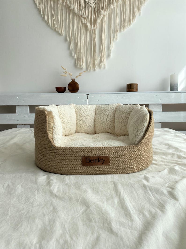 May include: A dog bed with a woven, light brown exterior and a soft, cream-colored interior. The bed has a rectangular shape with raised sides and a small, brown leather tag that reads "Bentley".