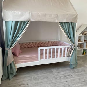 May include: A white wooden house-shaped bed frame featuring a beige fabric roof canopy, sage green curtains with white pom-pom trim, a pink fitted sheet, a matching pink star-shaped pillow, and a braided pink fabric bed bumper along the interior.