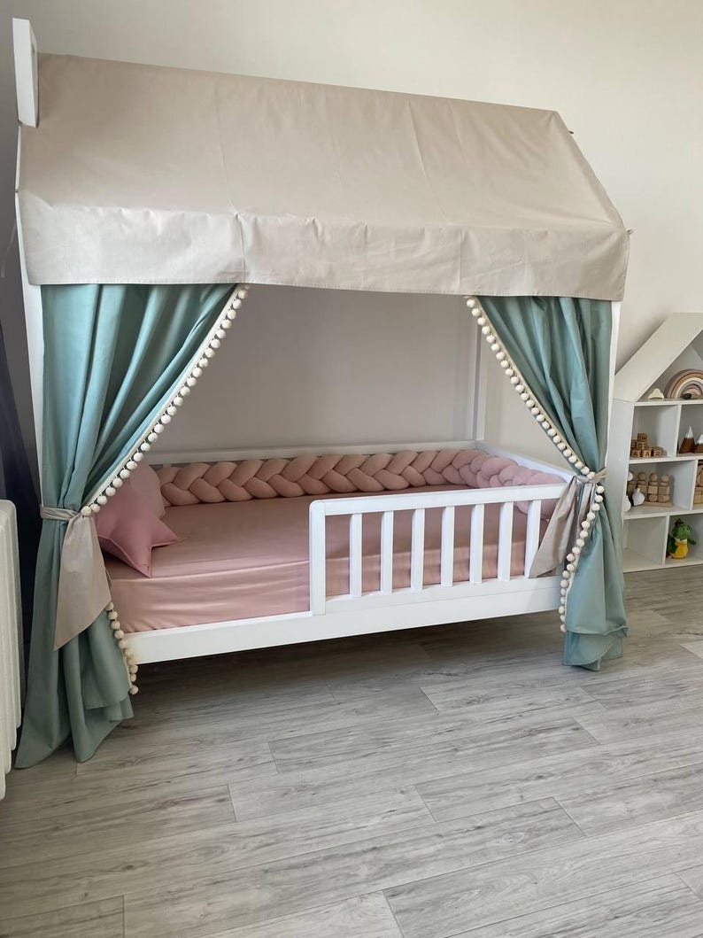 May include: A white wooden house-shaped bed frame featuring a beige fabric roof canopy, sage green curtains with white pom-pom trim, a pink fitted sheet, a matching pink star-shaped pillow, and a braided pink fabric bed bumper along the interior.