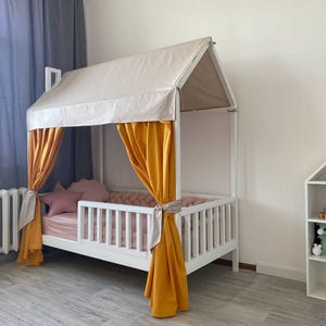 May include: A white house-shaped bed frame featuring a beige fabric roof canopy and mustard yellow curtains tied to the side posts. The bed includes a pink mattress, a pink star-shaped pillow, and a braided pink bumper along the interior rails.