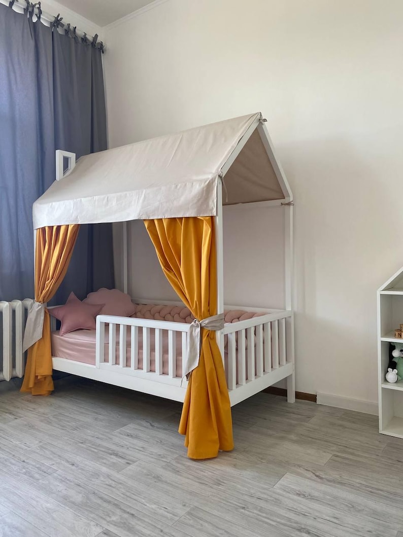 May include: A white house-shaped bed frame featuring a beige fabric roof canopy and mustard yellow curtains tied to the side posts. The bed includes a pink mattress, a pink star-shaped pillow, and a braided pink bumper along the interior rails.