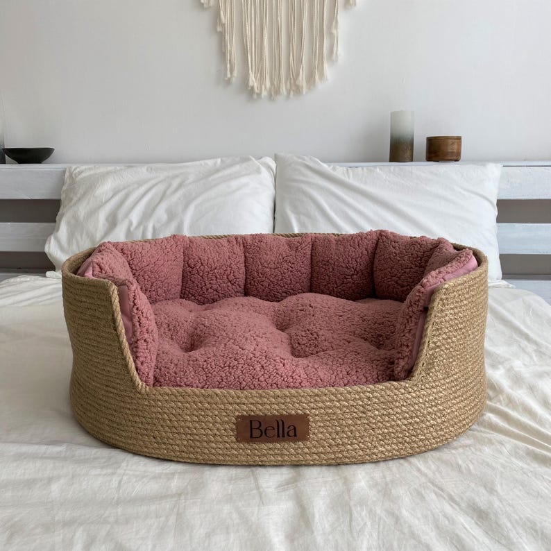 May include: A round pet bed with a woven, natural-colored exterior and a plush, rose-colored interior. The bed has a small, rectangular tag that reads "Bella". The bed is designed for comfort and is suitable for small to medium-sized pets.
