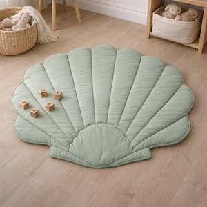Könnte beinhalten: Eine muschelförmige Spielmatte in Salbeigrün mit Steppdetails. Die Matte liegt auf einem hellen Holzboden, mit Holzbuchstabenwürfeln darauf. Der Raum ist in neutralen Tönen gehalten, mit einem kleinen Holztisch und Korb.