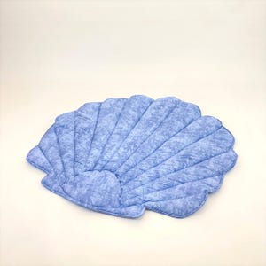 May include: A light blue, seashell-shaped cushion with a quilted pattern. The cushion has a scalloped edge and a textured, marbled appearance. The design resembles a large, open seashell.