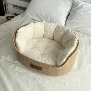 May include: A beige pet bed with a woven exterior and a soft, cream-colored interior. The bed has a rounded shape with a low rim and a stitched cushion. A small brown tag with the name "Bentley" is attached to the front.