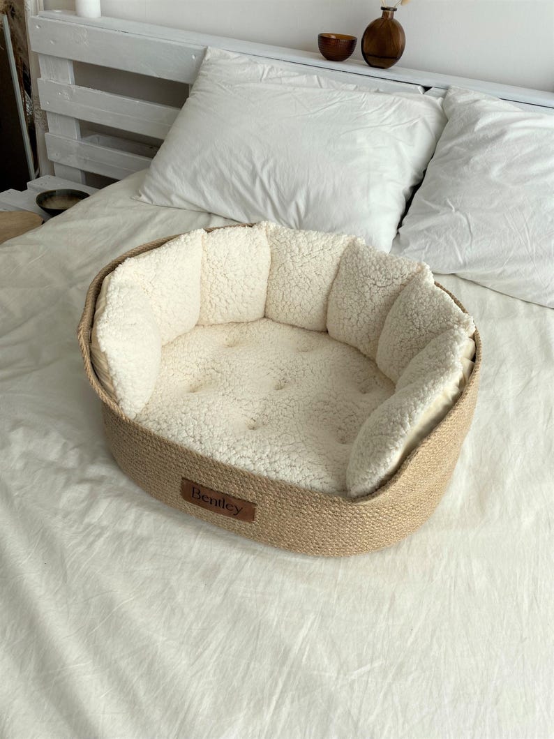 May include: A beige pet bed with a woven exterior and a soft, cream-colored interior. The bed has a rounded shape with a low rim and a stitched cushion. A small brown tag with the name "Bentley" is attached to the front.