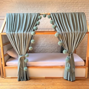 May include: A wooden bed frame with a canopy of sage green curtains and pom-pom accents. The bed has a white mattress and is set against a white brick wall. The curtains are tied back with matching fabric.