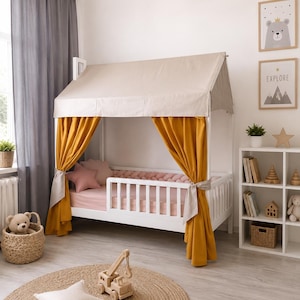 May include: A white wooden house-shaped bed frame featuring a beige fabric roof canopy and mustard yellow curtains tied to the side posts. The bed includes a pink mattress, a pink braided bumper, and a pink star-shaped pillow.