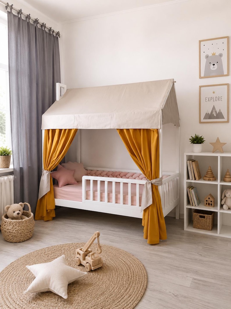 May include: A white wooden house-shaped bed frame featuring a beige fabric roof canopy and mustard yellow curtains tied to the side posts. The bed includes a pink mattress, a pink braided bumper, and a pink star-shaped pillow.