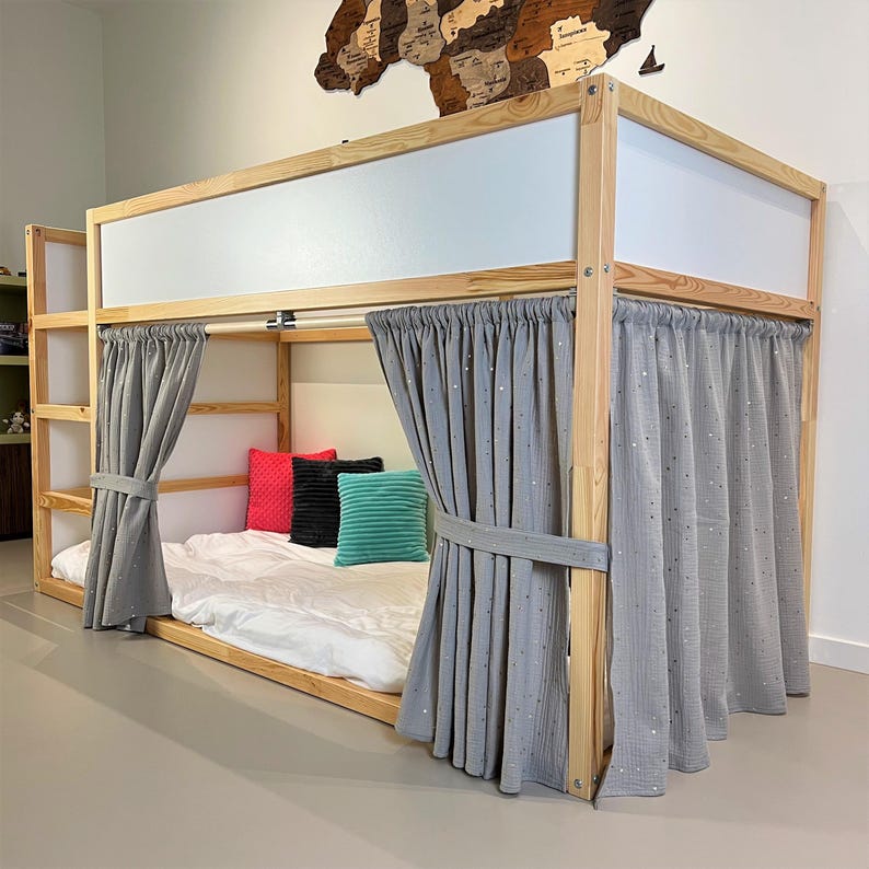 May include: A wooden bunk bed with a white mattress, gray curtains, and colorful pillows. The bed frame is made of light-colored wood. The curtains are tied back with matching fabric. The pillows are red, black, and teal.