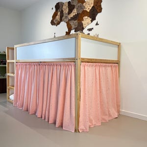 May include: A wooden bunk bed with a light pink curtain. The bed frame is made of light-colored wood. A wooden map of Ukraine hangs on the white wall above the bed.