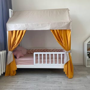 May include: A white wooden house-shaped bed frame featuring a beige fabric roof canopy, mustard yellow side curtains tied back, and a pink mattress with a matching pink star-shaped pillow and a braided pink bumper guard along the interior edge.